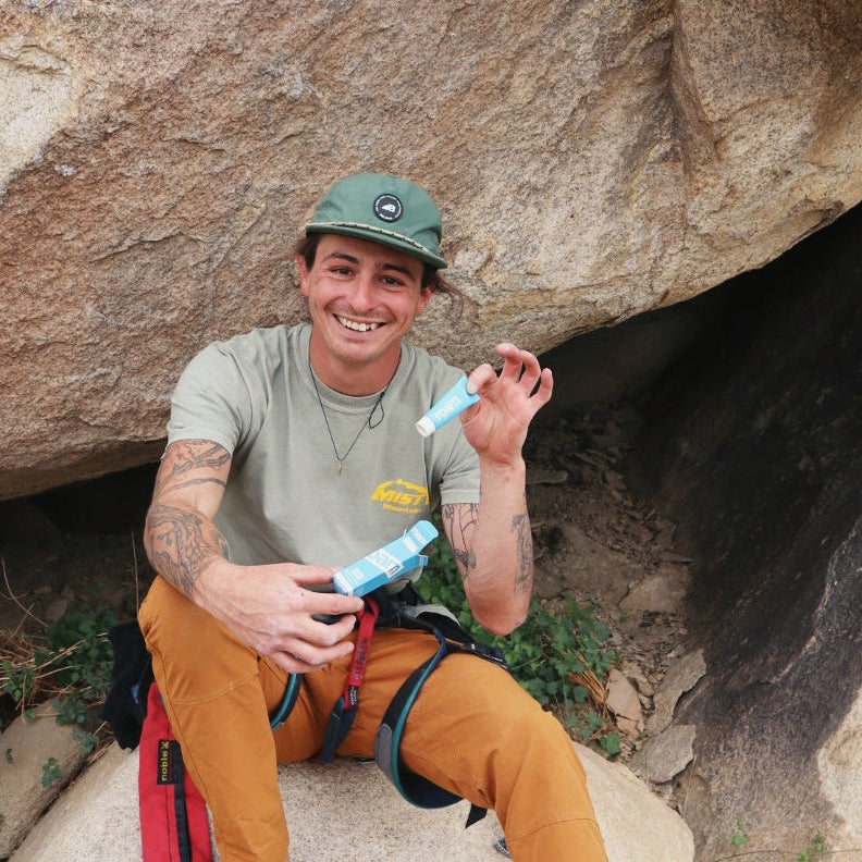 Person sitting on a rock with climbing gear, holding a tube of roam unscented deodorant.